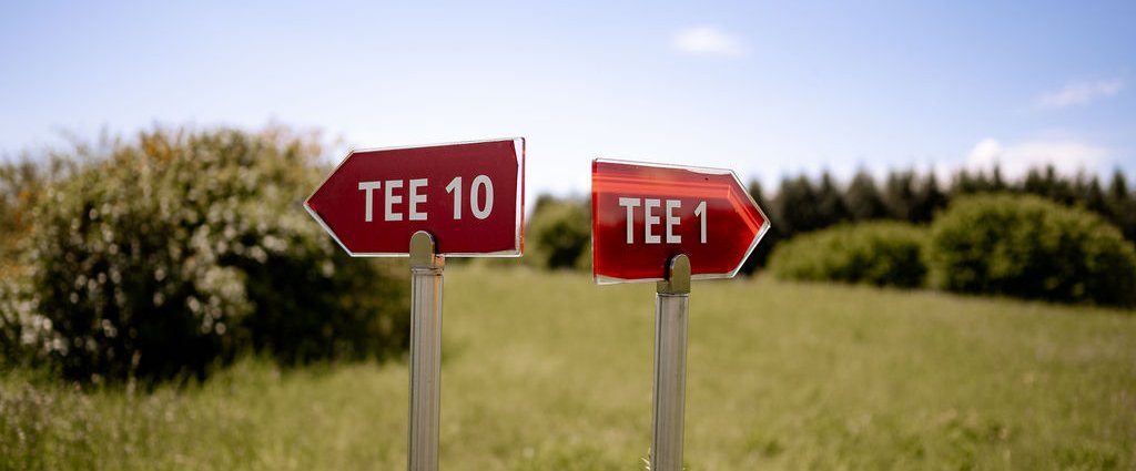 Auf einem Golfplatz stehende rote Wegweiser mit den Aufschriften ‚Tee 1‘ und ‚Tee 10‘.