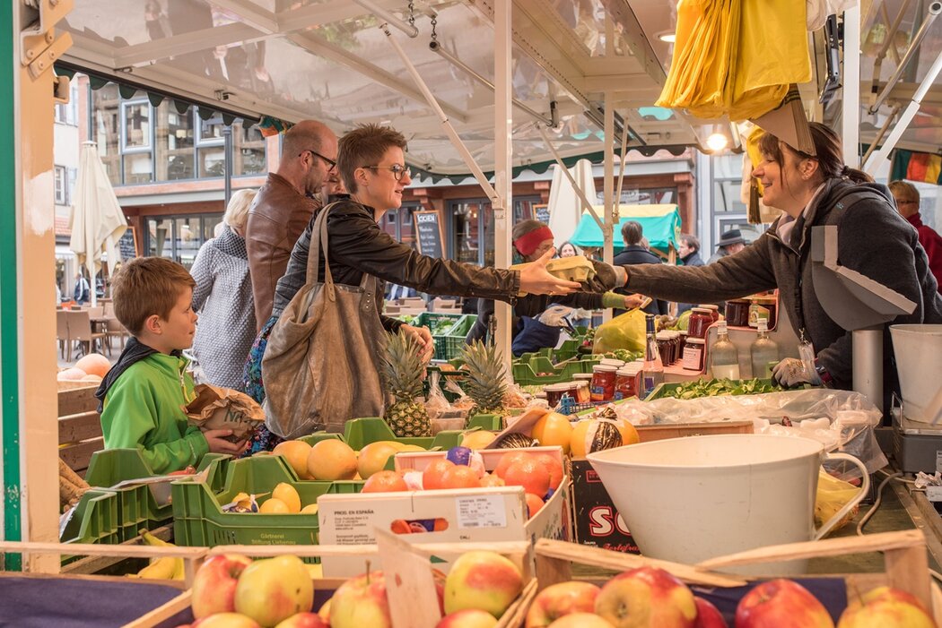 Eine lebhafte Szene auf dem Wochenmarkt in Bad Saulgau. Eine Frau in einer Lederjacke kauft Waren bei einer Verkäuferin und hält dabei eine wiederverwendbare Tasche in der Hand. Neben ihr steht ein Junge, der eine Papiertüte hält. Der Verkaufsstand ist gefüllt mit frischen Produkten wie Äpfeln, Ananas und anderem Obst. Die Verkäuferin lächelt, während sie die Waren überreicht. Im Hintergrund sind weitere Käufer und bunte Marktstände zu sehen.