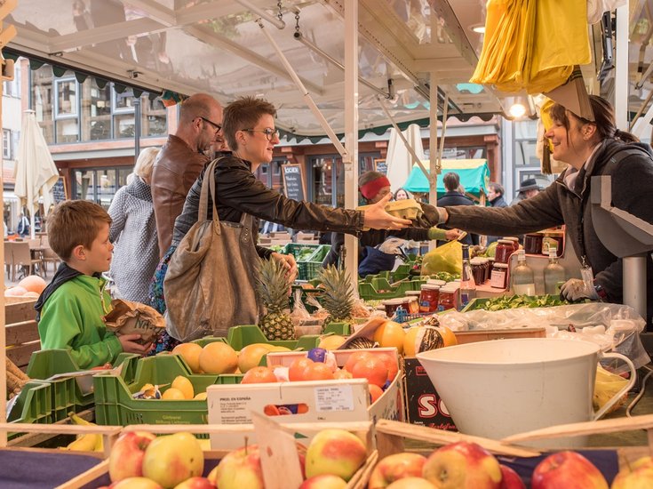 Eine lebhafte Szene auf dem Wochenmarkt in Bad Saulgau. Eine Frau in einer Lederjacke kauft Waren bei einer Verkäuferin und hält dabei eine wiederverwendbare Tasche in der Hand. Neben ihr steht ein Junge, der eine Papiertüte hält. Der Verkaufsstand ist gefüllt mit frischen Produkten wie Äpfeln, Ananas und anderem Obst. Die Verkäuferin lächelt, während sie die Waren überreicht. Im Hintergrund sind weitere Käufer und bunte Marktstände zu sehen.
