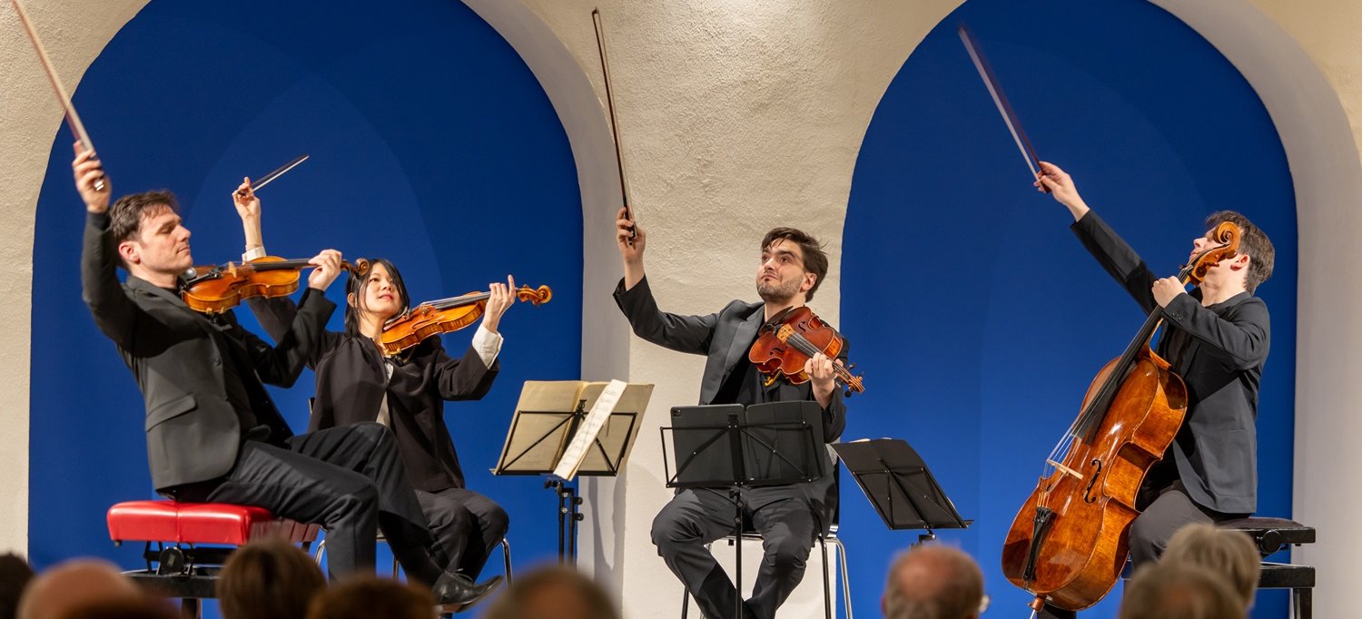 Streichquartett mit zwei Geigern, einem Bratschisten und einem Cellisten spielt vor Publikum in einem Raum mit weißen Rundbögen vor blauer Wand.