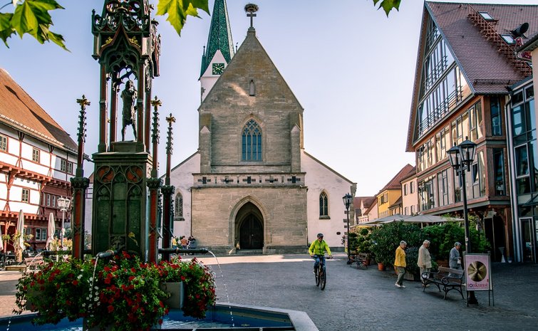 Historischer Marktplatz von Bad Saulgau mit gotischer Kirche, einem verzierten Brunnen mit Blumen sowie traditionellen und modernen Gebäuden; Menschen flanieren und ein Radfahrer fährt vorbei, bei sonnigem, Herbst-Wetter.